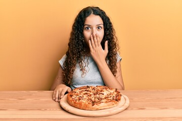 Teenager hispanic girl eating italian pizza covering mouth with hand, shocked and afraid for...