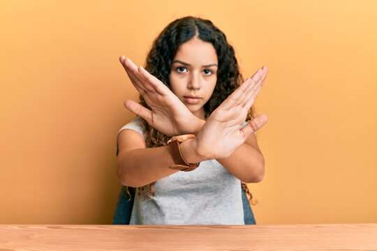 Teenager hispanic girl wearing casual clothes sitting on the table rejection expression crossing arms and palms doing negative sign, angry face