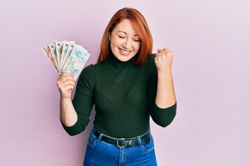 Beautiful redhead woman holding united kingdom pounds screaming proud, celebrating victory and...