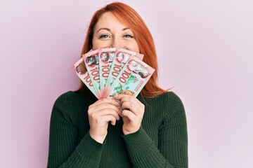 Beautiful redhead woman holding 100 new zealand dollars banknote smiling with a happy and cool smile on face. showing teeth.