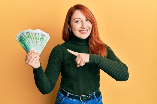 Beautiful redhead woman holding 5 singapore dollars banknotes smiling happy pointing with hand and finger