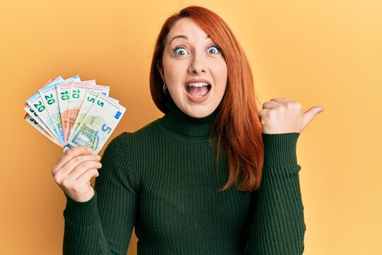 Beautiful redhead woman holding euro banknotes pointing thumb up to the side smiling happy with open mouth