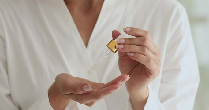 Close up young woman in bathrobe holding bottle, applying hyaluronic collagen vital vitamin C serum intense drops on hands, enjoying skincare routine after morning showering, prevent skin ageing.