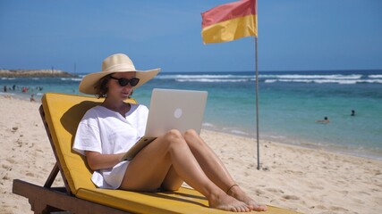 Digital nomad concept. Caucasian female in a straw hat works with a laptop on a beach