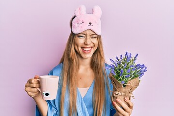 Beautiful young blonde woman wearing pajama drinking a cup of infused lavender winking looking at...