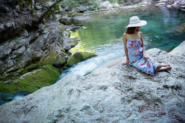 Young caucasian woman in a dress and hat resting on stones near a mountain river.