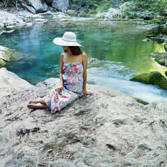 Young caucasian woman in a dress and hat resting on stones near a mountain river.