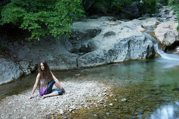 Young slim caucasian woman with long brown hair doing yoga in the mountains near a mountain river.