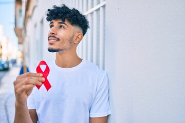 Young arab man smiling happy holding hiv awaraness red ribbon leaning on the wall.