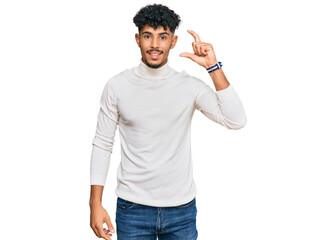 Young arab man wearing casual winter sweater smiling and confident gesturing with hand doing small size sign with fingers looking and the camera. measure concept.