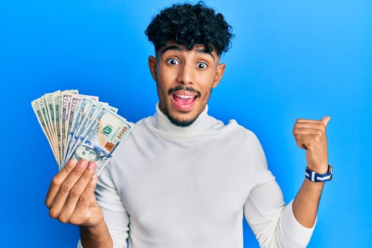 Young arab handsome man holding dollars pointing thumb up to the side smiling happy with open mouth