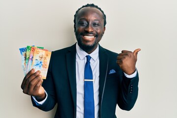 Handsome young black man wearing business suit holding franc swiss banknotes pointing thumb up to the side smiling happy with open mouth