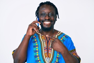 Young african american man with braids wearing ethnic tshirt having conversation talking on the smartphone smiling happy pointing with hand and finger