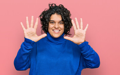 Young hispanic woman with curly hair wearing turtleneck sweater showing and pointing up with fingers number ten while smiling confident and happy.