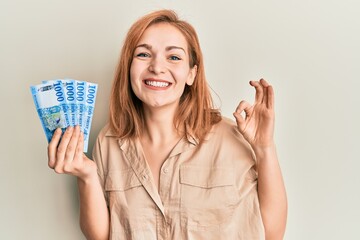 Obraz premium Young caucasian woman holding hungarian forint banknotes doing ok sign with fingers, smiling friendly gesturing excellent symbol