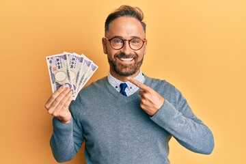 Handsome middle age man holding 5000 japanese yen banknotes smiling happy pointing with hand and finger