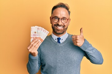 Handsome middle age man holding 10 colombian pesos banknotes smiling happy and positive, thumb up doing excellent and approval sign