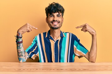 Young hispanic man wearing casual clothes sitting on the table looking confident with smile on...