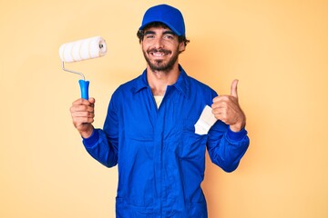 Handsome young man with curly hair and bear wearing builder jumpsuit uniform holding paint roller smiling happy and positive, thumb up doing excellent and approval sign