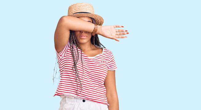 Young african american woman with braids wearing summer hat covering eyes with arm, looking serious and sad. sightless, hiding and rejection concept