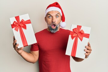 Middle age handsome man wearing christmas hat and holding a gifts afraid and shocked with surprise and amazed expression, fear and excited face.