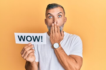 Middle age handsome man holding wow word on paper covering mouth with hand, shocked and afraid for...