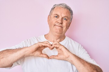 Senior grey-haired man wearing casual white tshirt smiling in love showing heart symbol and shape with hands. romantic concept.