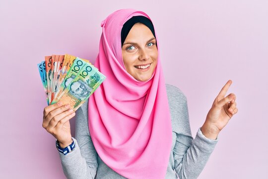 Young caucasian woman wearing islamic hijab holding australian dollars banknotes smiling happy pointing with hand and finger to the side