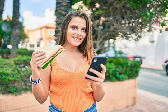 Young middle east girl smiling happy using smartphone and eating sandwich at the city.