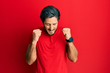 Young hispanic man wearing casual red t shirt excited for success with arms raised and eyes closed...
