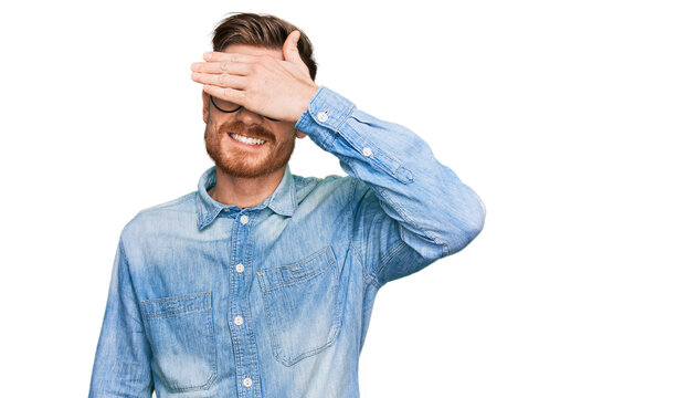 Young redhead man wearing casual denim shirt smiling and laughing with hand on face covering eyes for surprise. blind concept.