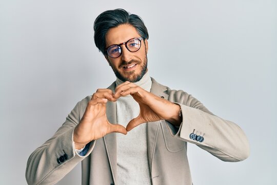 Young hispanic man wearing business jacket and glasses smiling in love doing heart symbol shape with hands. romantic concept.