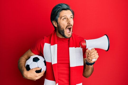 Young hispanic man football hooligan holding ball using megaphone angry and mad screaming frustrated and furious, shouting with anger looking up.