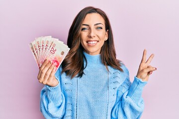 Young brunette woman holding 20 israel shekels banknotes smiling looking to the camera showing...