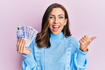 Young brunette woman holding 20 swedish krona banknotes pointing thumb up to the side smiling happy with open mouth