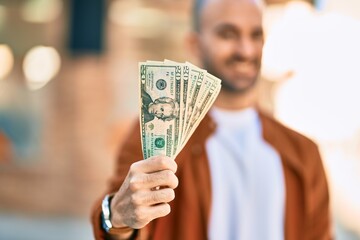 Young hispanic bald man smiling happy holding usa dollars at the city.