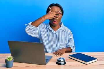 Young african woman working at hotel reception using laptop peeking in shock covering face and eyes with hand, looking through fingers with embarrassed expression.