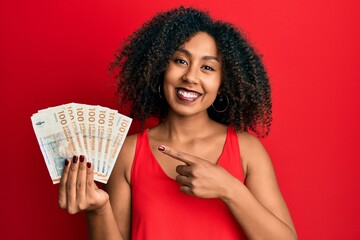 Beautiful african american woman with afro hair holding 100 danish krone banknotes smiling happy...