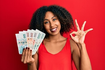 Beautiful african american woman with afro hair holding czech koruna banknotes doing ok sign with fingers, smiling friendly gesturing excellent symbol