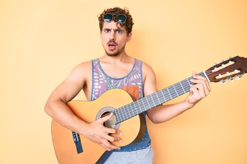 Young caucasian man with curly hair playing classical guitar in shock face, looking skeptical and...