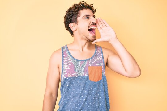 Young caucasian man with curly hair wearing casual style with sleeveless shirt shouting and screaming loud to side with hand on mouth. communication concept.
