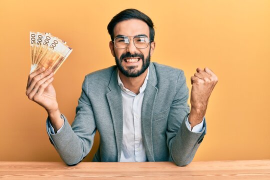 Young hispanic man holding norwegian krone banknotes sitting on the table screaming proud, celebrating victory and success very excited with raised arm