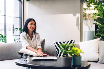 Smiling happy beautiful balck business woman wearing white shirt relaxing using laptop.Young hipster girl freelancer working and sitting on sofa in green plant terrace decorated office