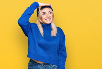 Young caucasian woman wearing wool winter sweater and cap very happy and smiling looking far away with hand over head. searching concept.