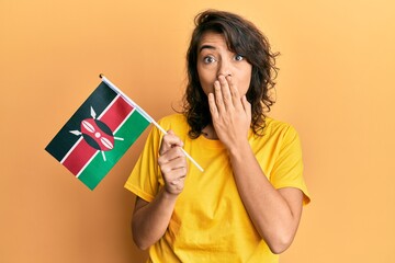 Young hispanic woman holding kenya flag covering mouth with hand, shocked and afraid for mistake....