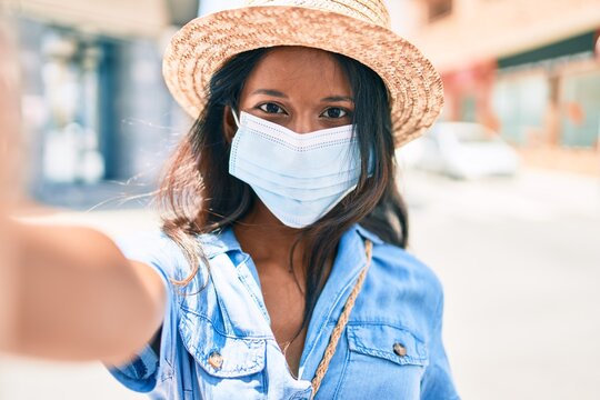 Young beautiful indian woman wearing summer hat and medical mask making selfie by the camera at the city.