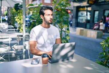Young hispanic man with serious expression working using laptop at coffee shop terrace.