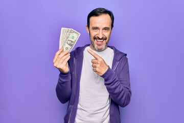Middle age handsome man holding bunch of dollars banknotes over isolated purple background smiling happy pointing with hand and finger