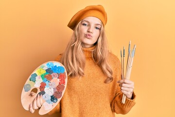 Beautiful young caucasian girl holding paintbrush and painter palette looking at the camera blowing a kiss being lovely and sexy. love expression.