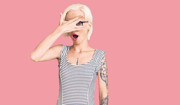 Young blonde woman with tattoo wearing casual clothes and glasses peeking in shock covering face and eyes with hand, looking through fingers with embarrassed expression.